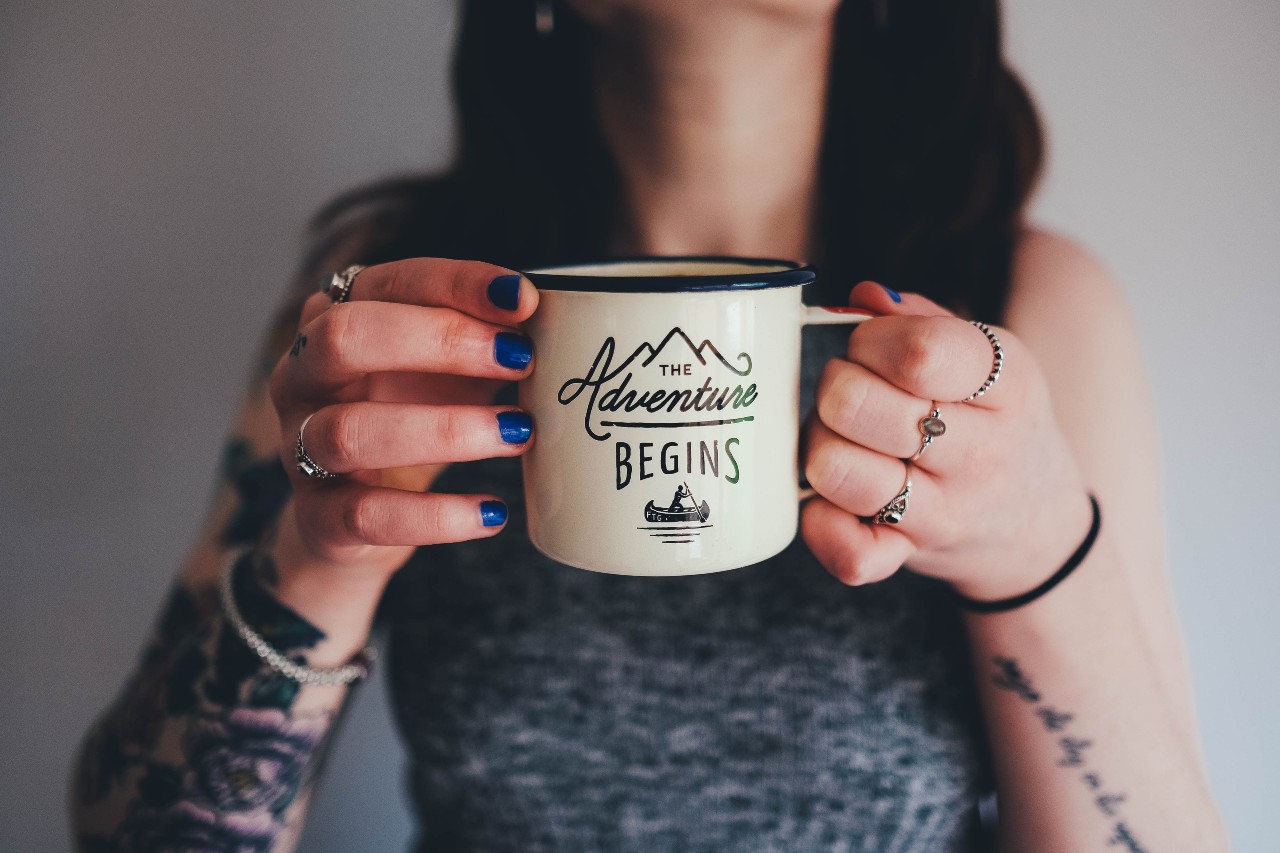A woman with blue nails and fashion rings sips coffee out of a mug that says “The Adventure Begins.”