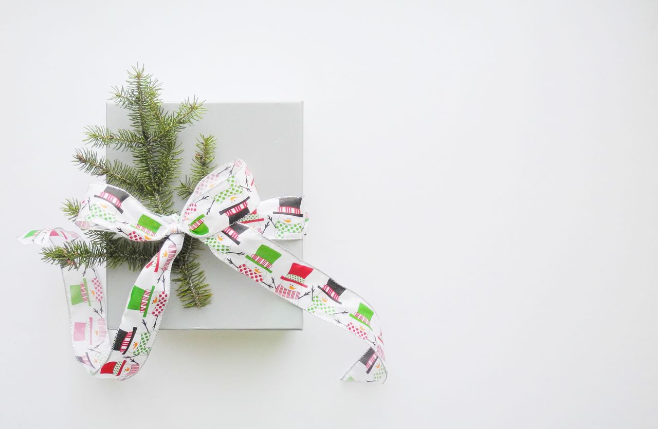 A wide view of a simple grey gift box on a grey backdrop, decorated with a snowman ribbon and a sprig of pine.