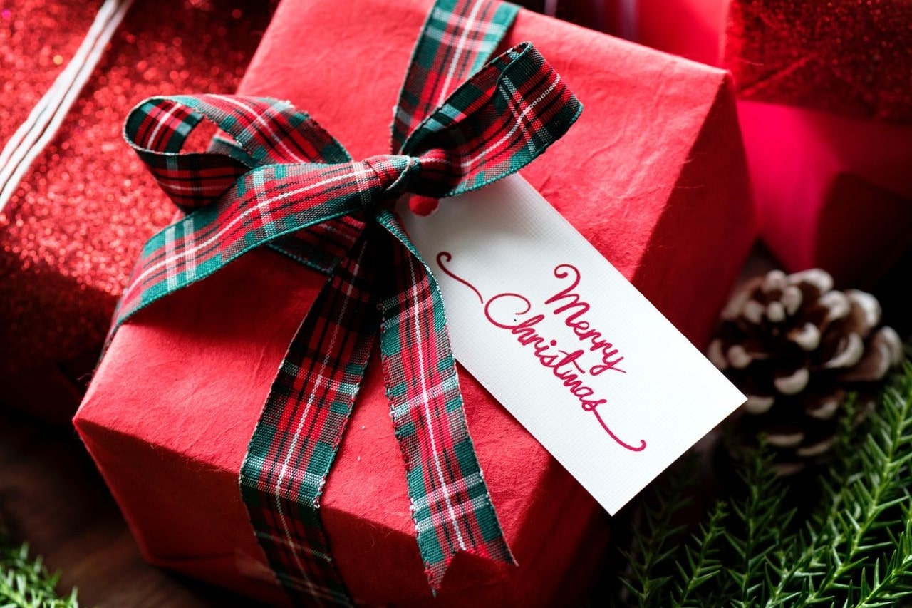 A close-up of a small red gift box adorned with a plaid ribbon nestled among a festive gift display, its tag reading “Merry Christmas.”