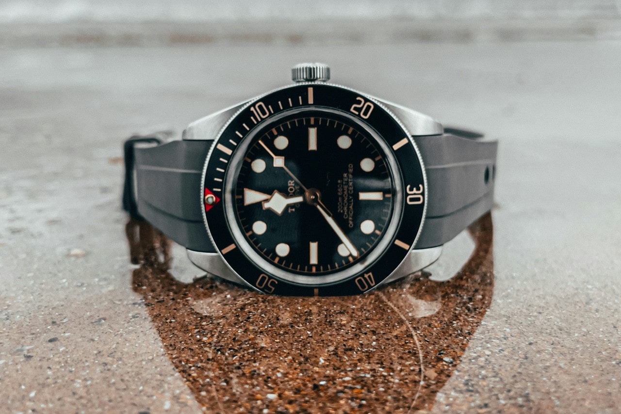 A TUDOR watch with a black dial and luminous markers rests on a wet, reflective surface.