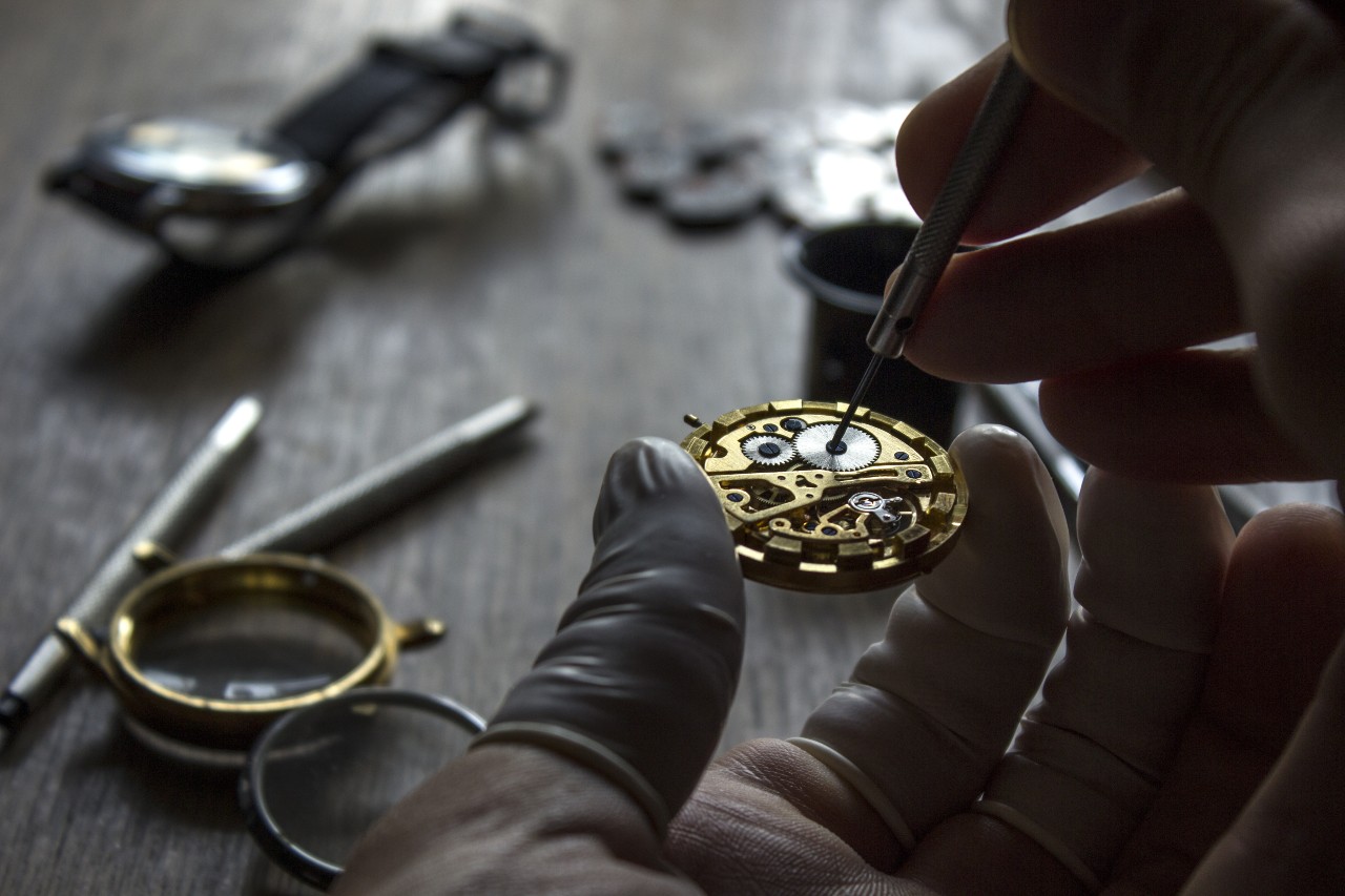 Close-up of a gloved hand delicately repairing a watch's intricate gears with a small tool.