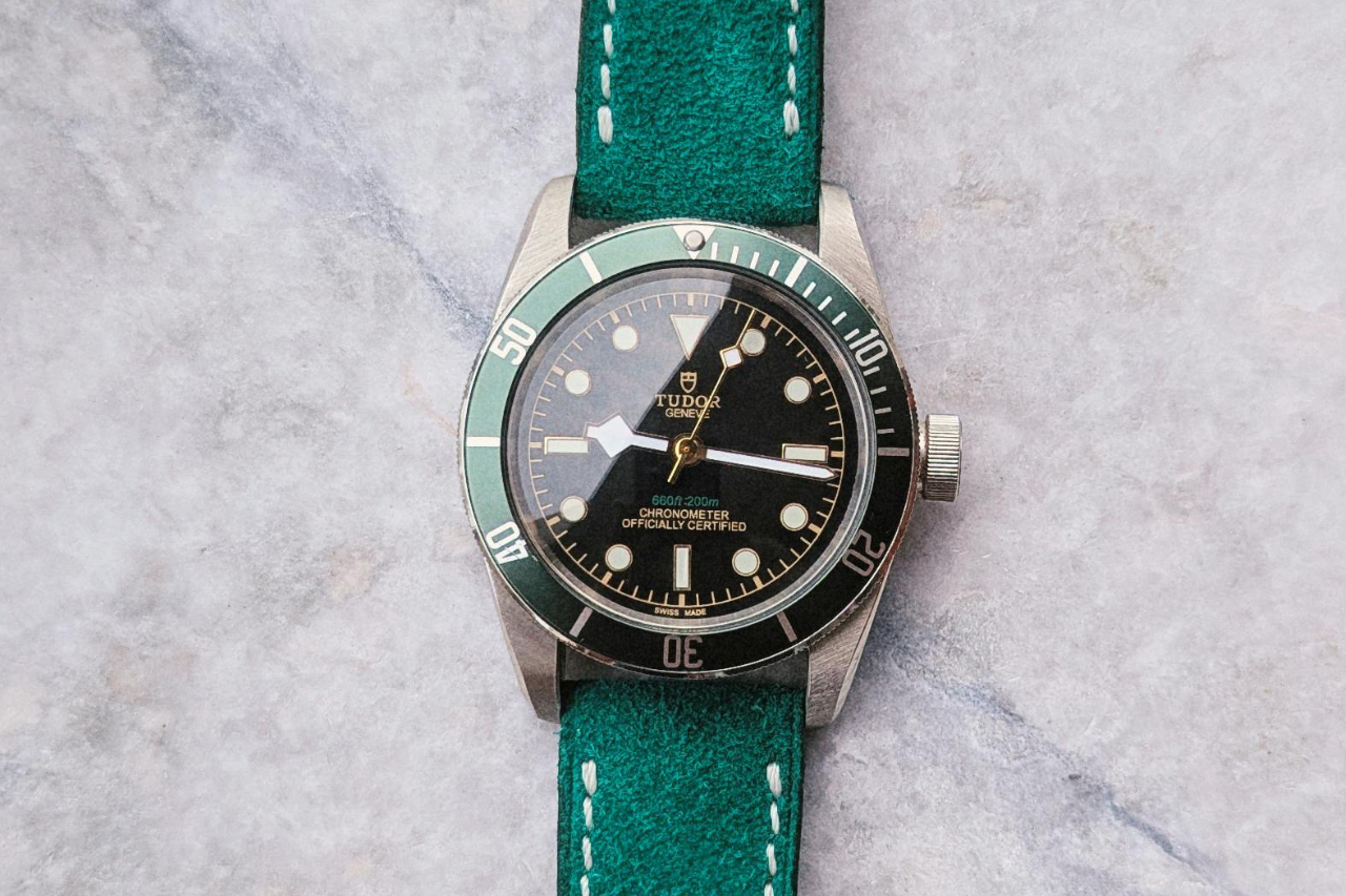 A vintage-style TUDOR wristwatch with a green bezel and a teal leather strap is placed on a light stone surface.