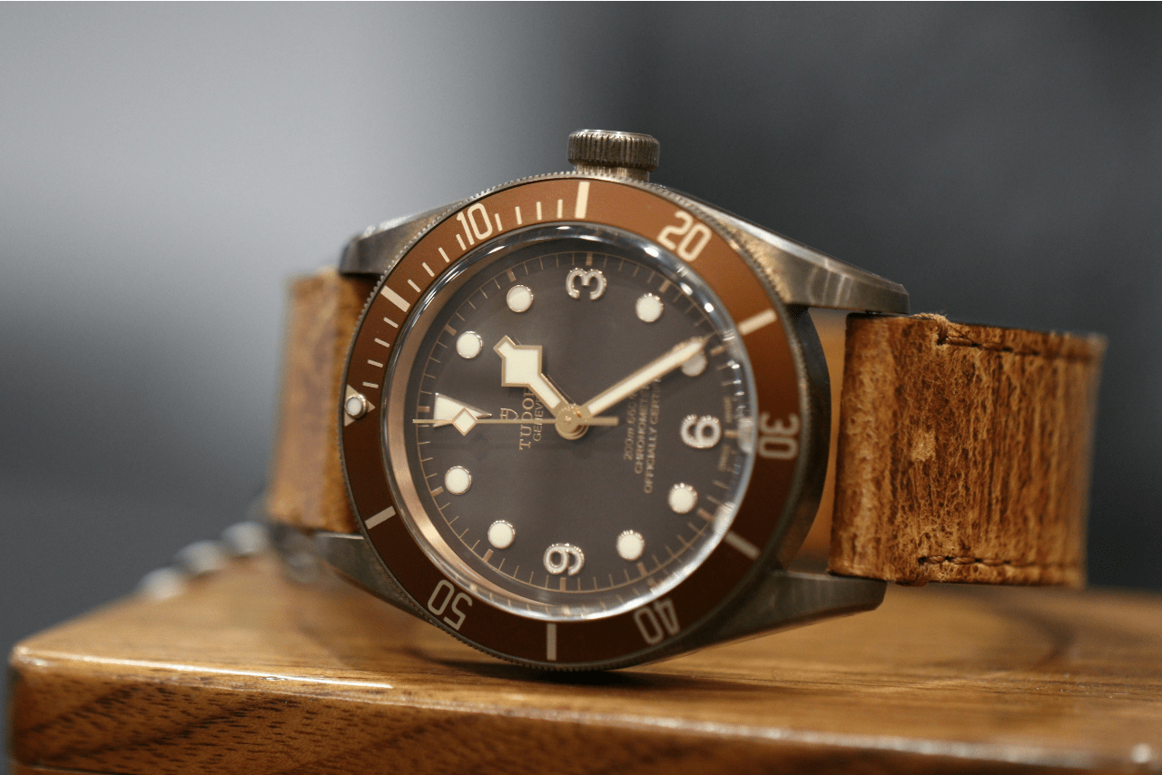 A TUDOR watch with a brown leather strap, bronze case, and a brown dial featuring white markers on a wooden surface.