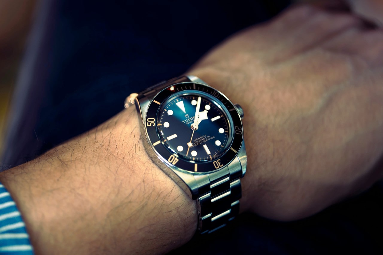 A close up of a TUDOR metallic wristwatch with a deep blue dial and 60 second rotating bezel on a man&rsquo;s hand.
