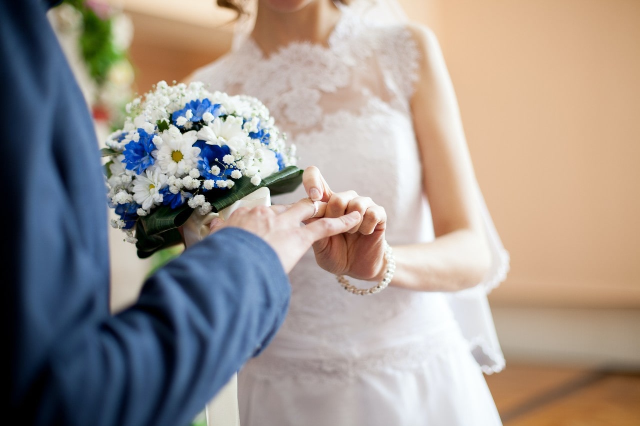 Close-up of a bride in a lace dress holding a blue and white bouquet while sliding a ring onto her groom's hand.