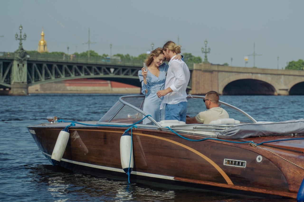 a couple standing on a small boat and embracing, the woman holding a flute of champagne