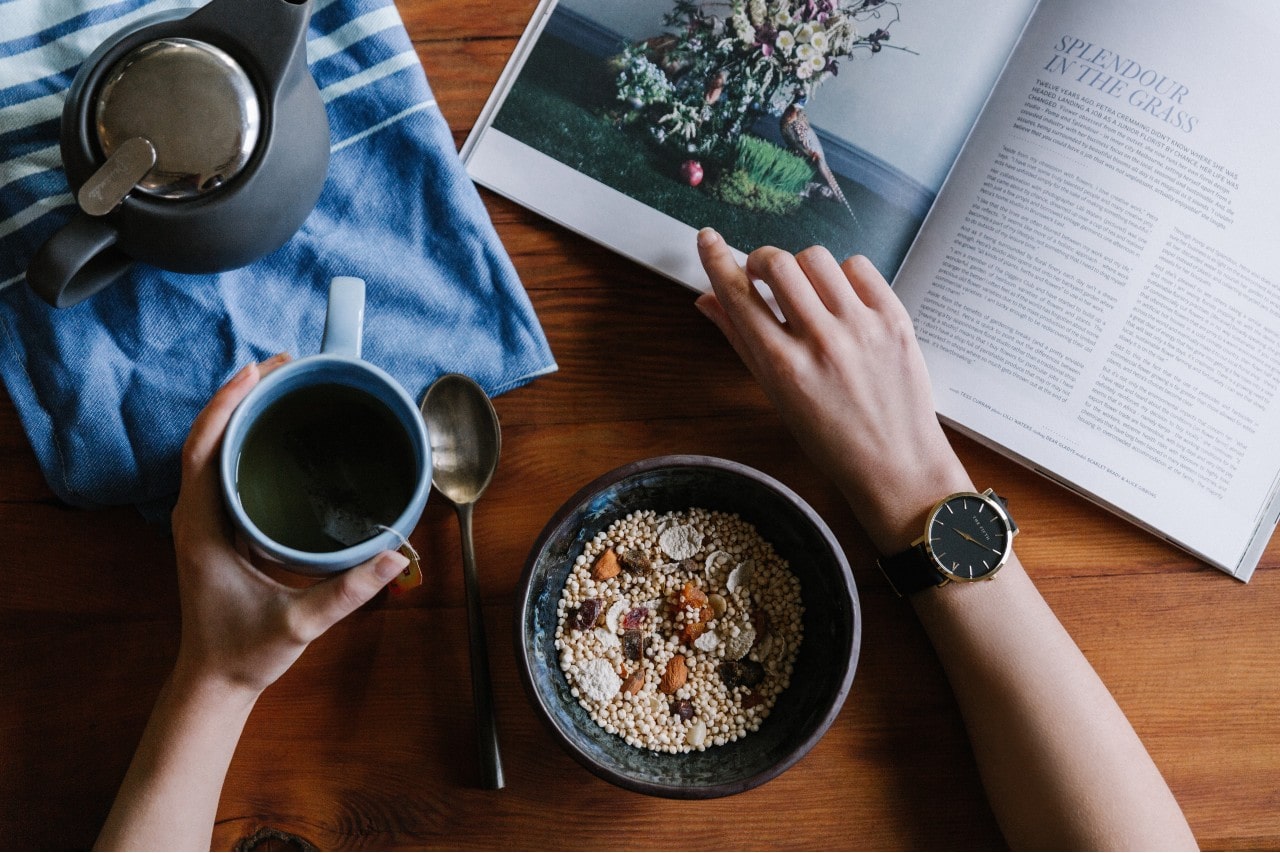 A man enjoying breakfast and coffee reads a magazine while wearing his watch.