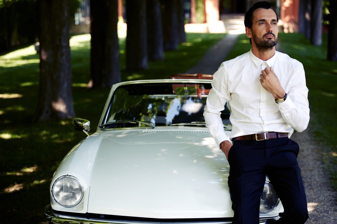 A man leans on a classic car while adjusting his collared shirt.