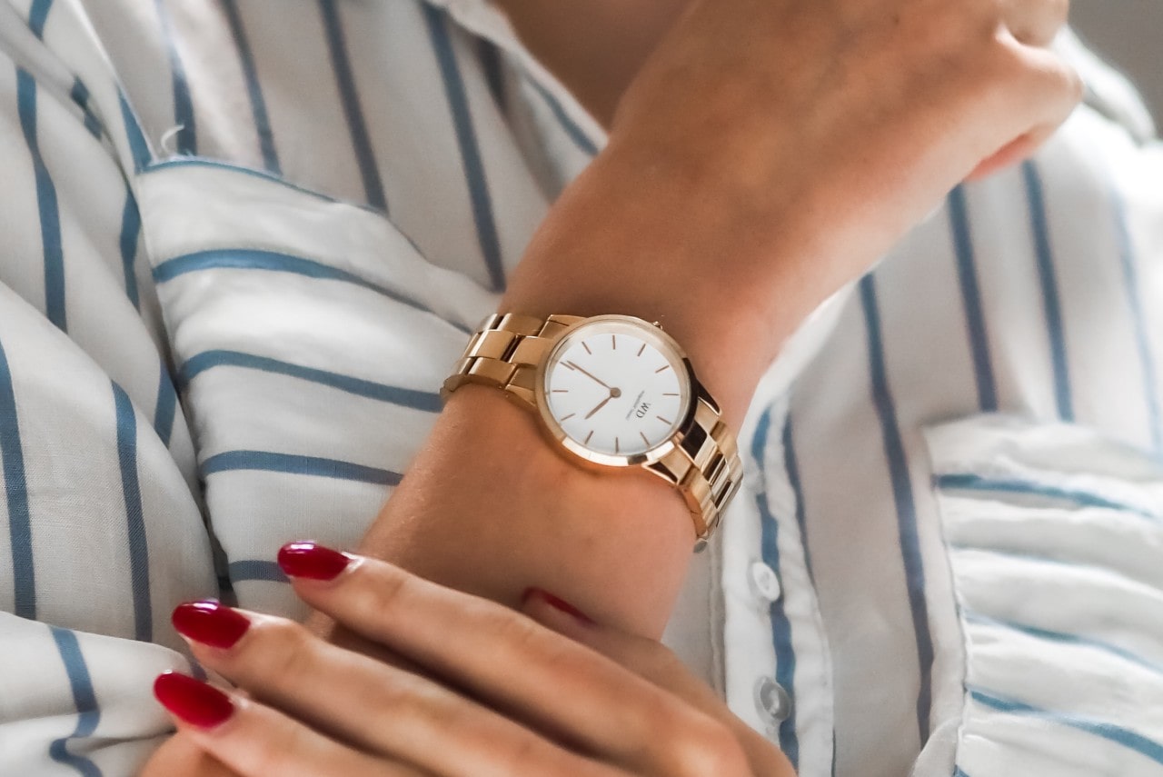 A woman with red manicured fingernails wears a golden watch with a white dial.