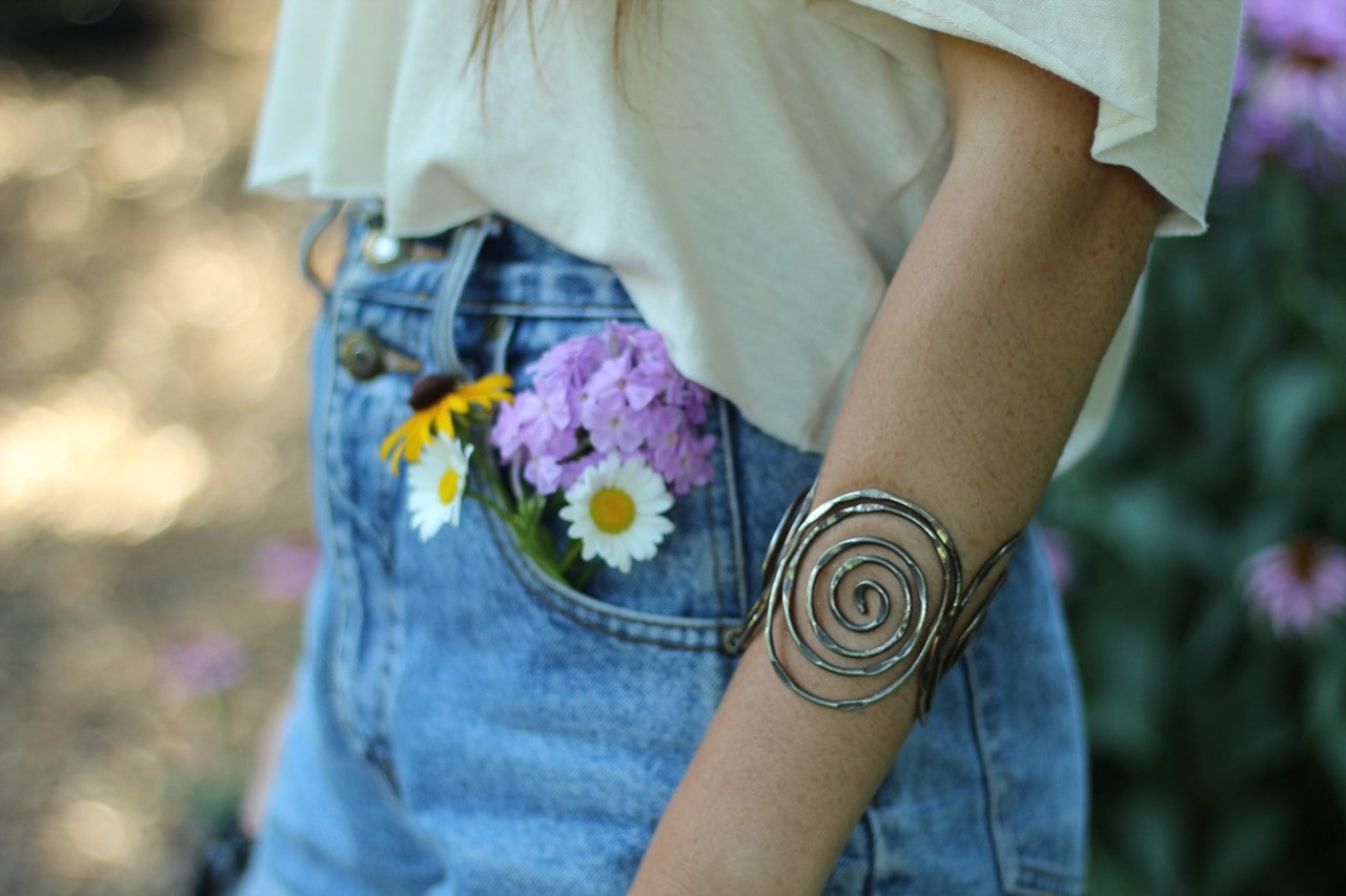 A woman wearing a silver swirled bangle stands in the forest with flowers tucked into her jeans.