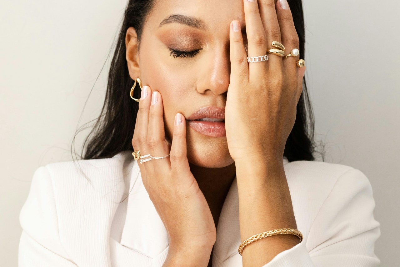 A woman adorned in yellow gold jewelry rests her hands on her face as she poses for the camera.