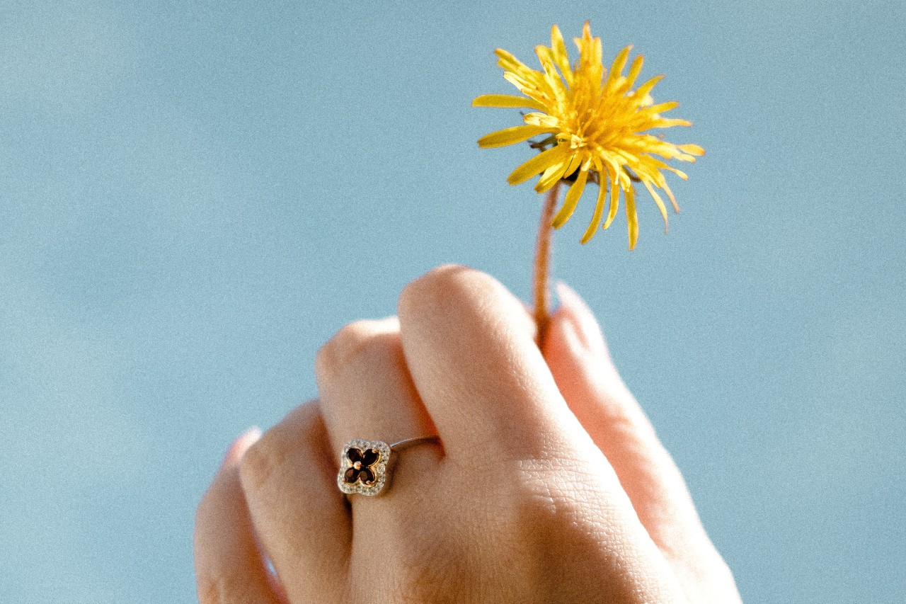 a hand holding a dandelion against a blue background and wearing a floral fashion ring
