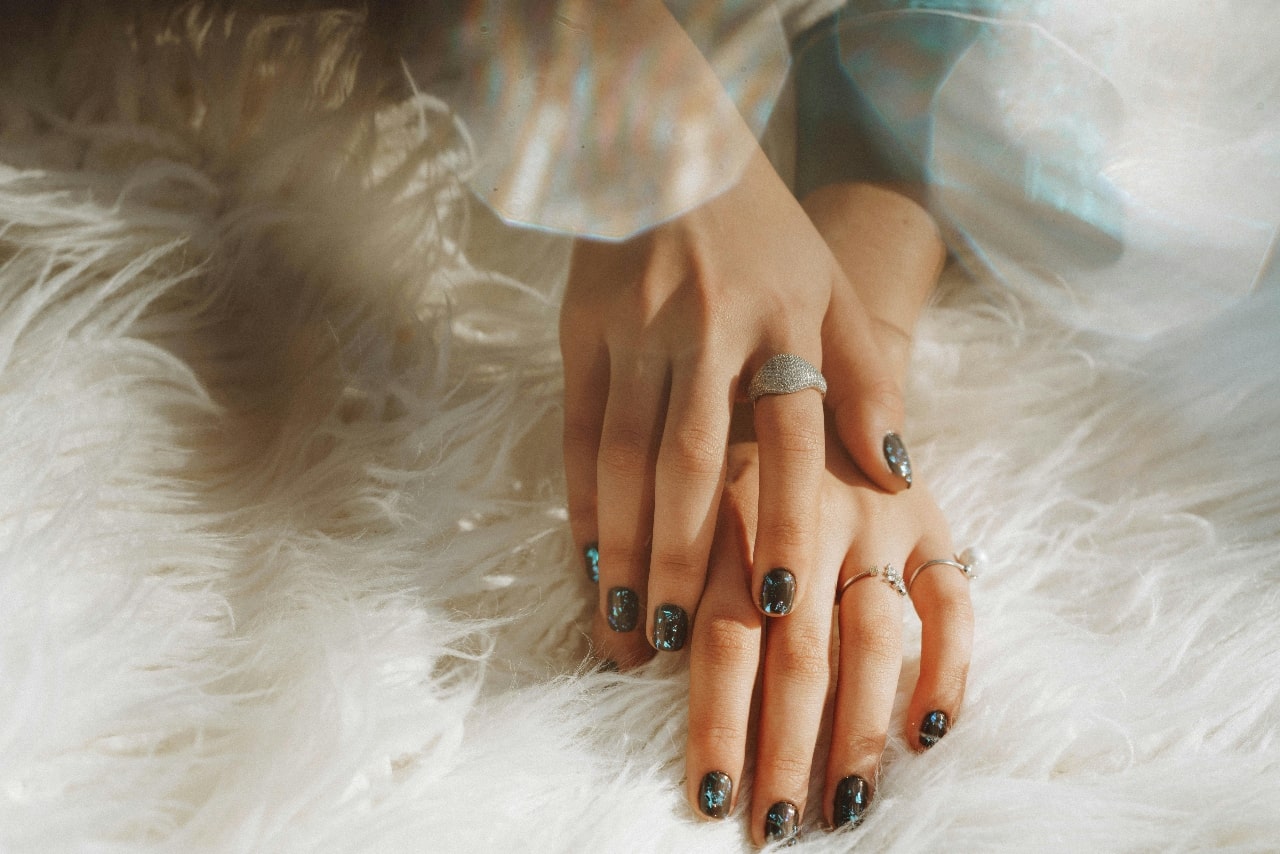 A woman’s hands with manicured nails on a white carpet, showcasing stackable rings and a diamond cocktail ring.