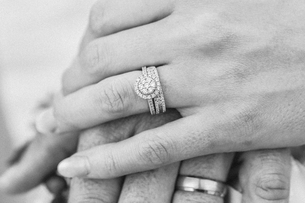 A close-up of two hands, showcasing a diamond engagement ring and wedding bands.