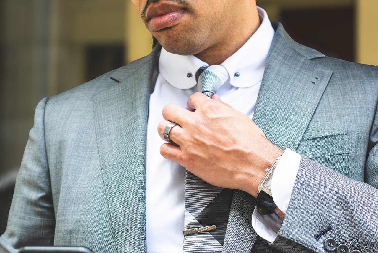 A close-up of a man in a grey suit fixing his tie with a tungsten wedding band and a leather wristwatch.