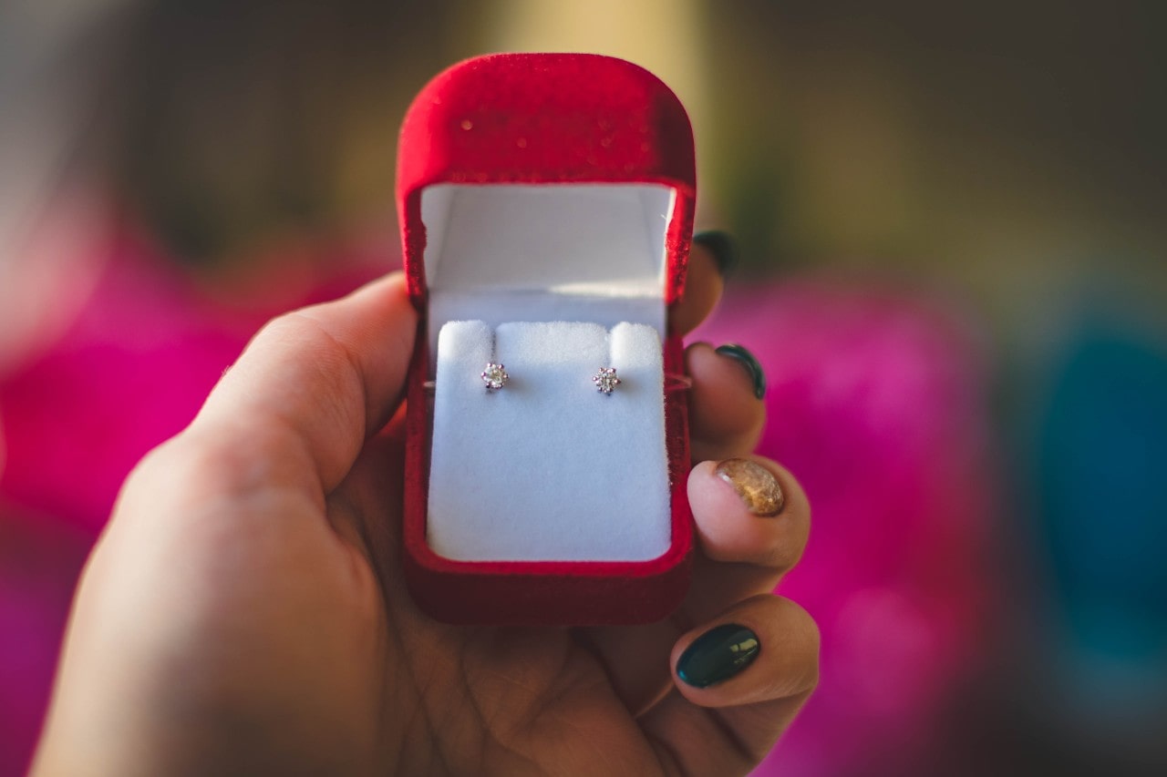 A nicely manicured hand holding a red velvet earring box with diamond studs inside.