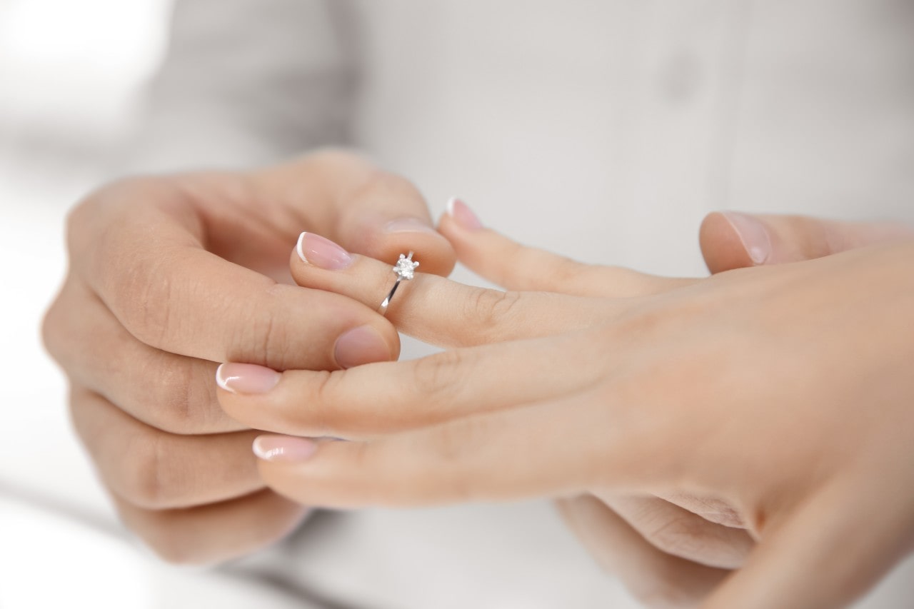 A person placing a solitaire diamond engagement ring on another’s finger.