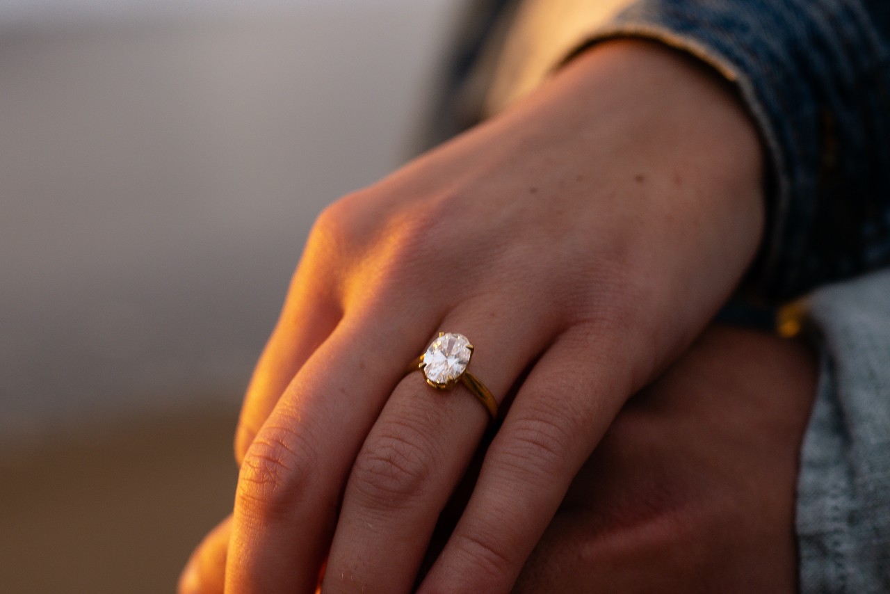 A gold engagement ring with a striking oval-cut diamond is shown on a hand in the glow of sunset light.