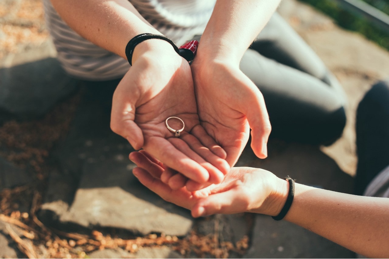 A person gently holds a solitaire engagement ring in their open hands, with another hand reaching out toward it.
