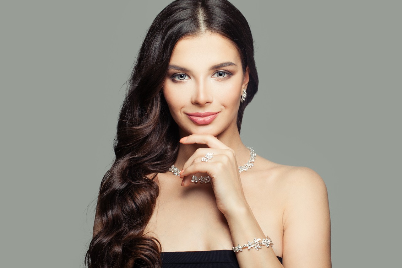 A woman with long dark hair poses against a neutral background, wearing a diamond necklace, matching earrings, bracelet, and rings, with her hand resting under her chin
