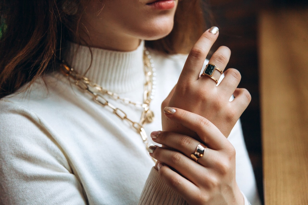 A woman in white turtleneck wearing yellow gold bulky chain necklaces and fashion rings