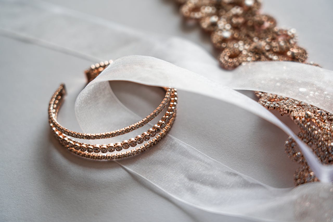 A close-up of rose gold diamond bangles displayed on a soft white fabric next to a rose gold ornamental design, a ribbon loosely draped around the bracelets.