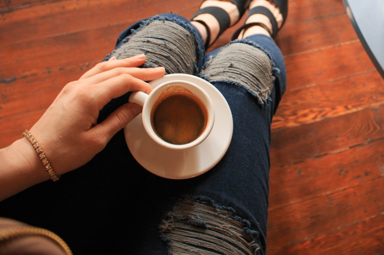 A top-down view of a woman wearing a rose gold tennis bracelet, ripped jeans and black sandals as she drinks a cup of coffee.