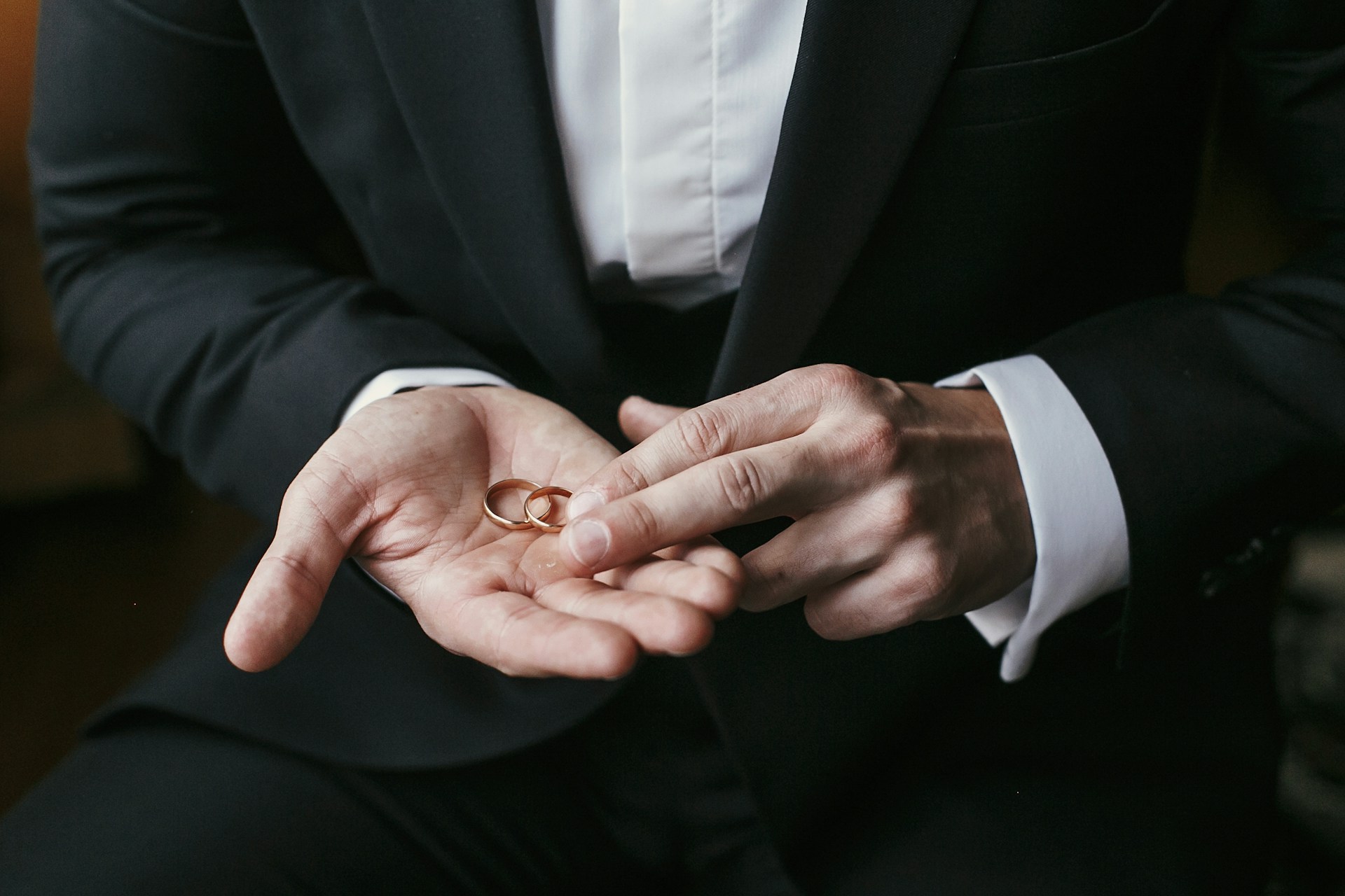 A close-up of a groom holding two gold wedding bands in the palm of his hand. A close-up of a groom holding two gold wedding bands in the palm of his hand.