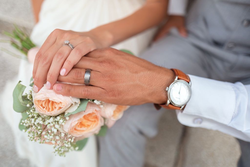 Close-up of a couple's hands with wedding bands, gently resting on a bouquet of peach roses and white flowers. Close-up of a couple's hands with wedding bands, gently resting on a bouquet of peach roses and white flowers.