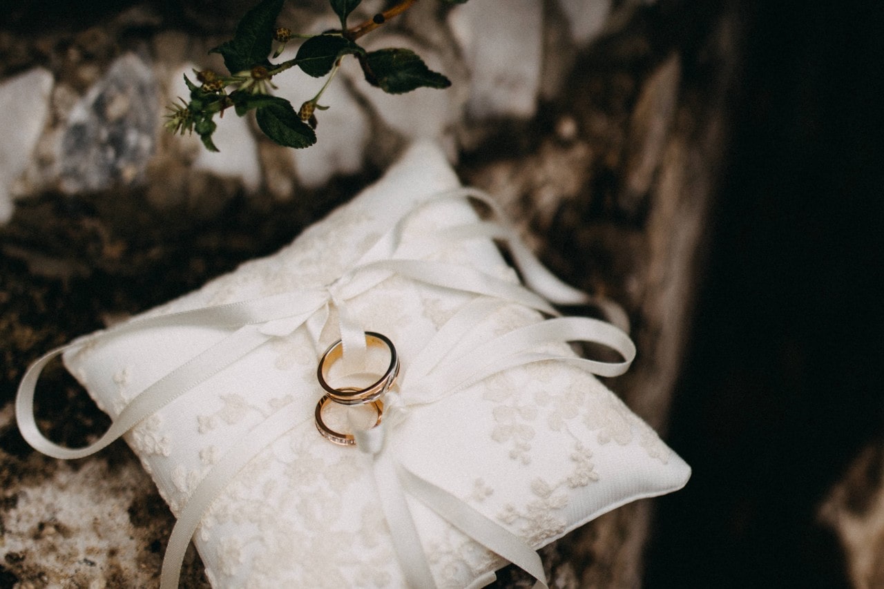 Two gold wedding rings resting on a white lace pillow with ribbons, set against a rustic stone background. Two gold wedding rings resting on a white lace pillow with ribbons, set against a rustic stone background.