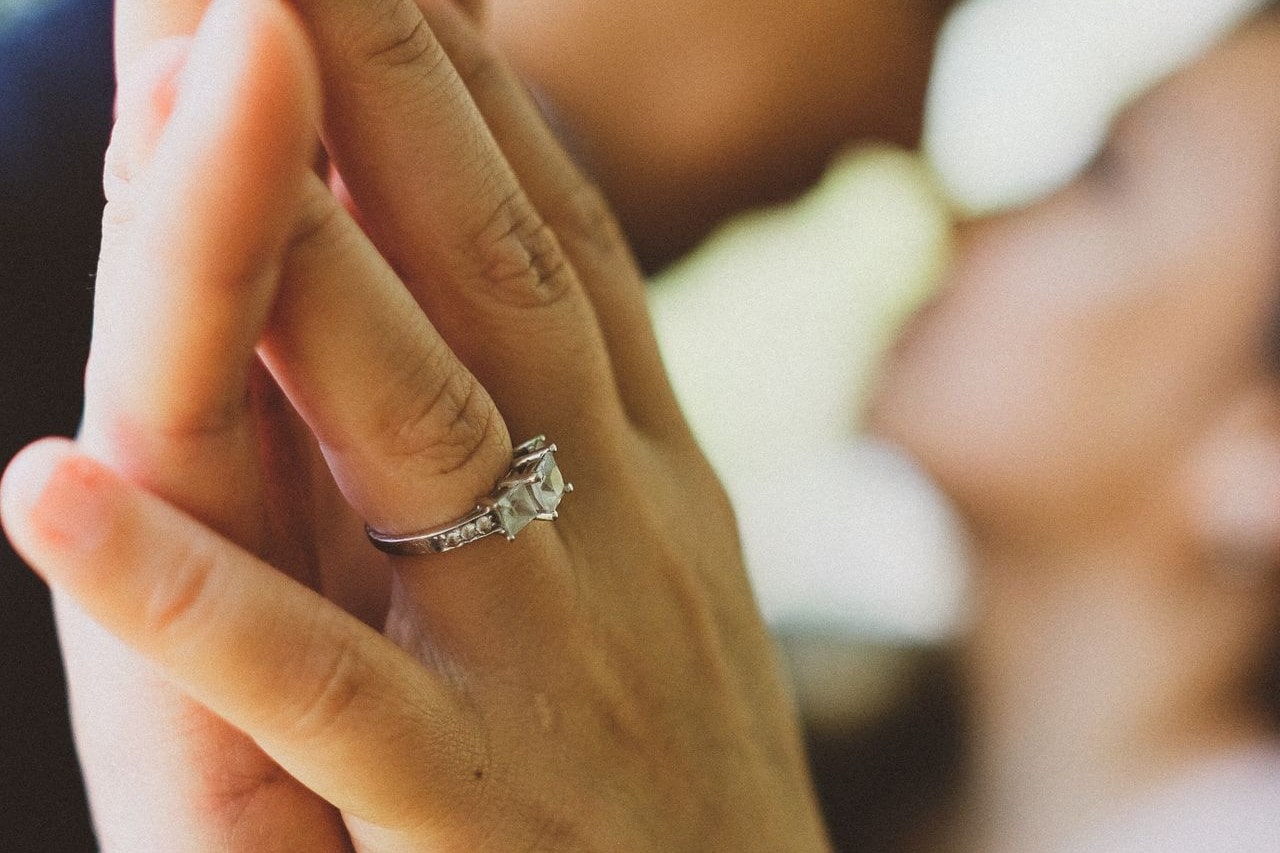 Close-up image of a couple’s hands pressed together, the woman wearing a princess cut engagement ring, their faces blurred in the background