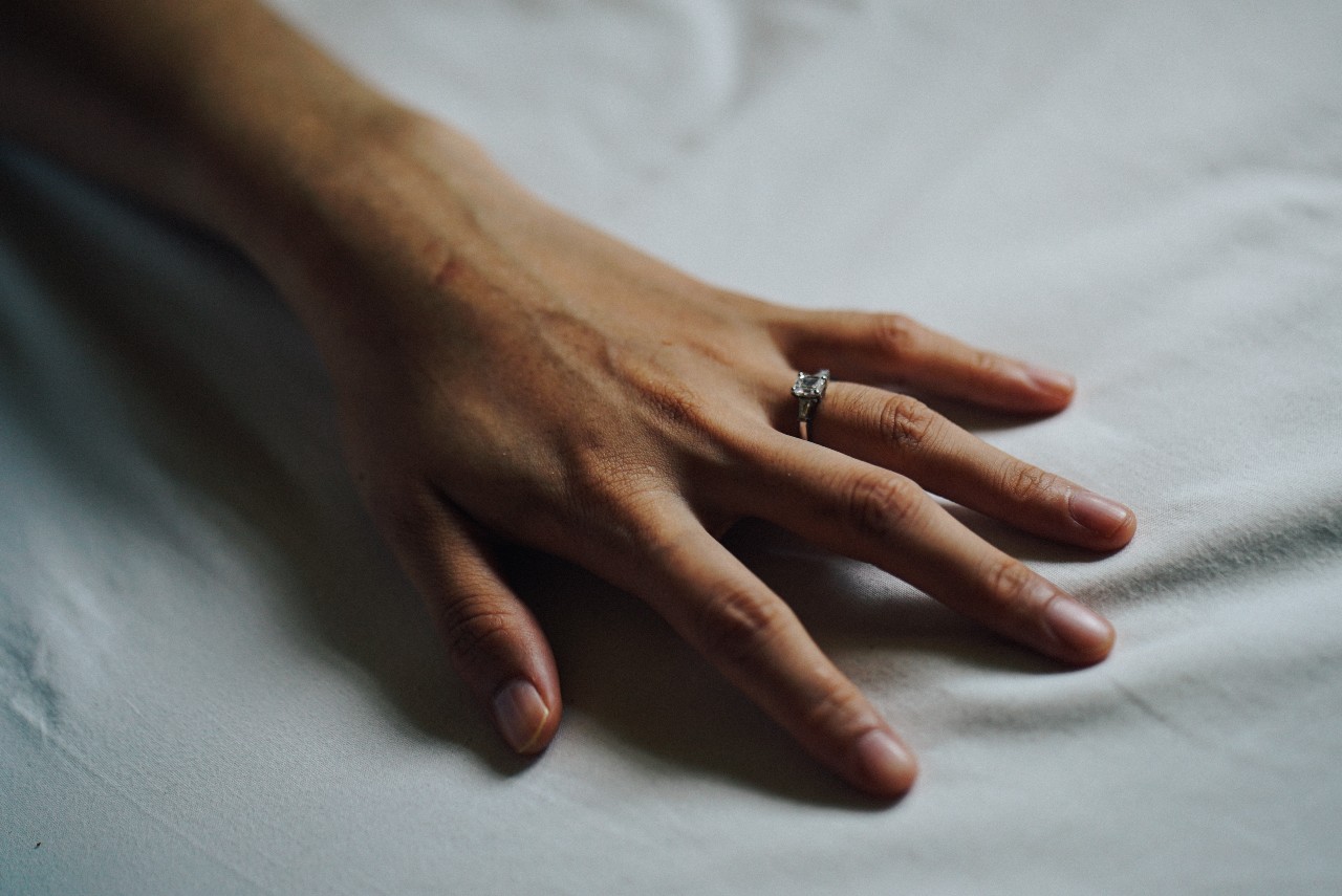 a hand lying on a white sheet, wearing a silver engagement ring with a princess cut center stone