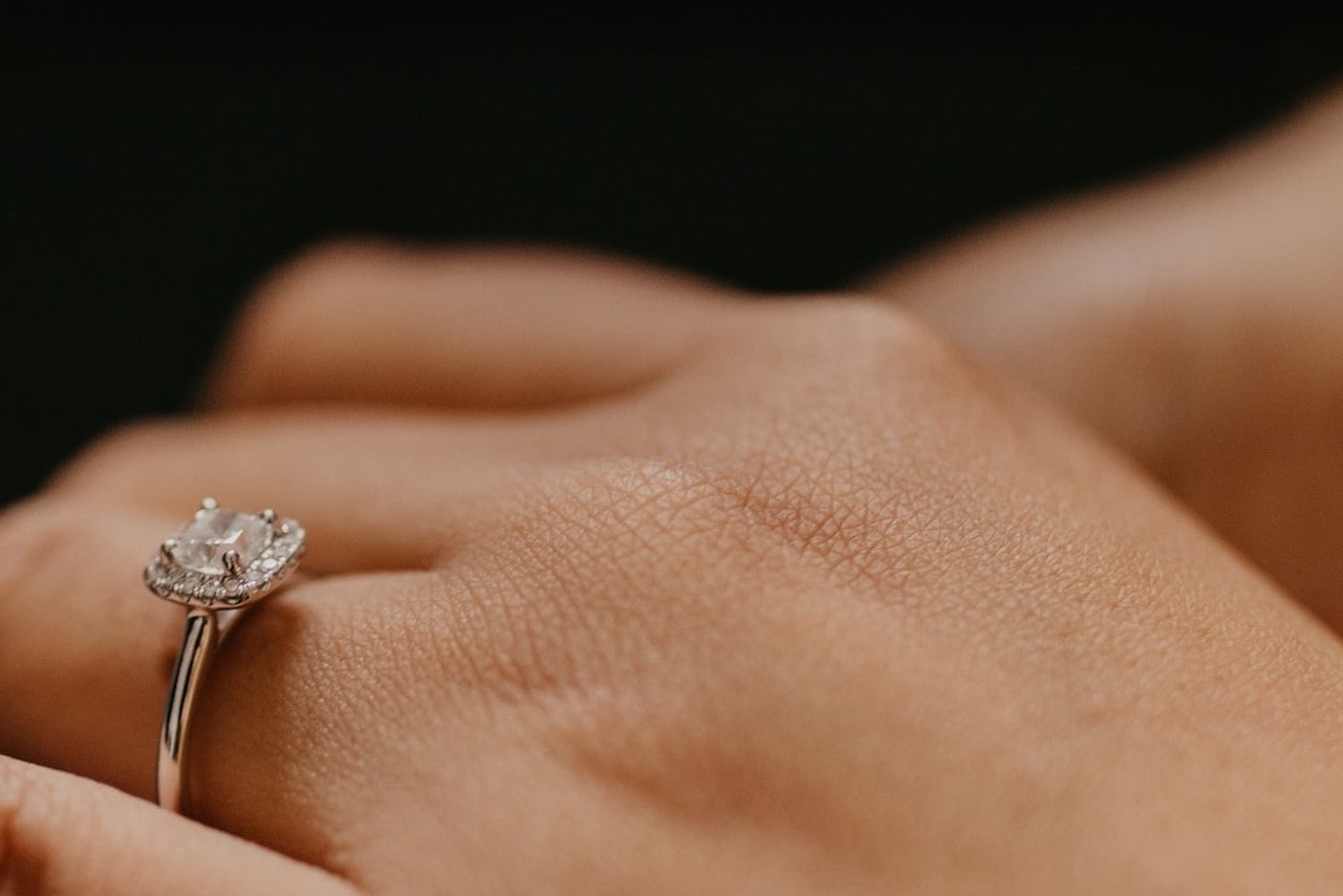 Close up image of a hand wearing a silver, princess cut engagement ring with a halo of accent stones