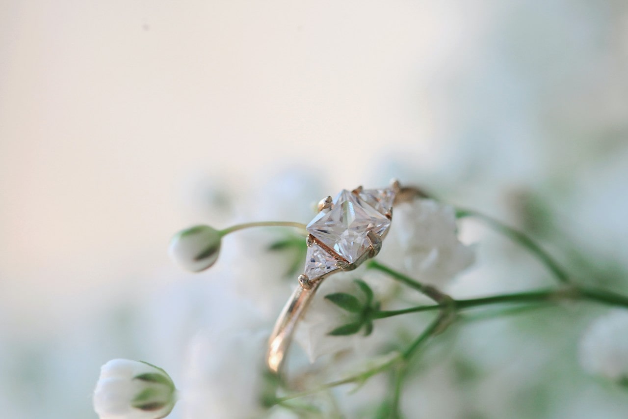 Close-up image of a three stone, gold engagement ring with a princess cut center stone on a bloom of flowers.