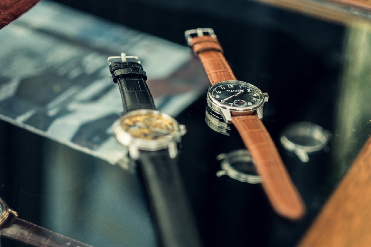 A close up of two leather wristwatches – one brown and the other black - on a black reflective surface.