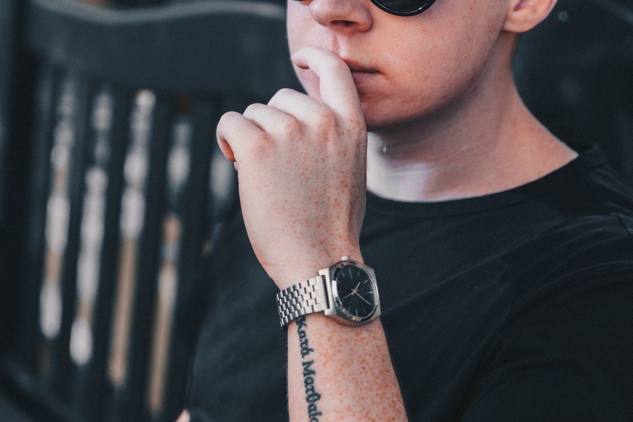 A man in a black t-shirt with tattoo on his hand, showcasing a metallic wristwatch with black dial.