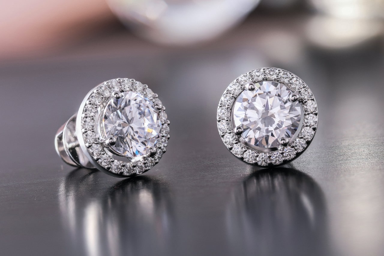 A pair of round halo diamond studs in white gold on a wooden surface A pair of round halo diamond studs in white gold on a wooden surface