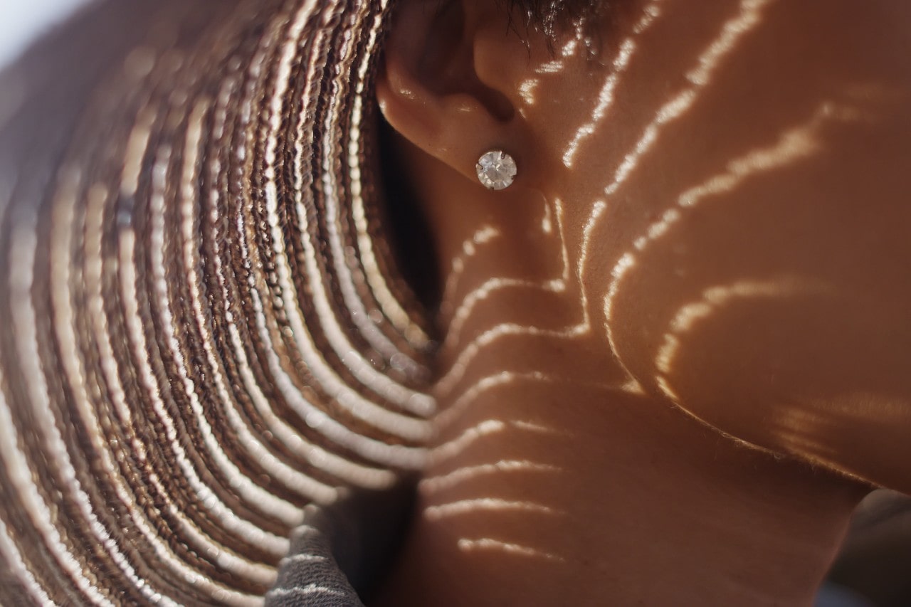 A close up of a woman wearing round diamond studs with striped sunlight patterns cast by a woven hat.
