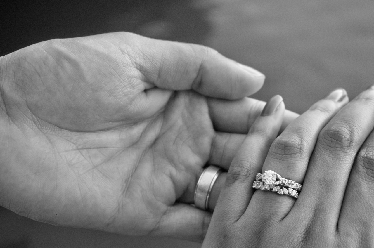 A close-up of a man’s hand holding a woman’s– his adorned with a wedding band, hers with a bridal stack. A close-up of a man’s hand holding a woman’s– his adorned with a wedding band, hers with a bridal stack.
