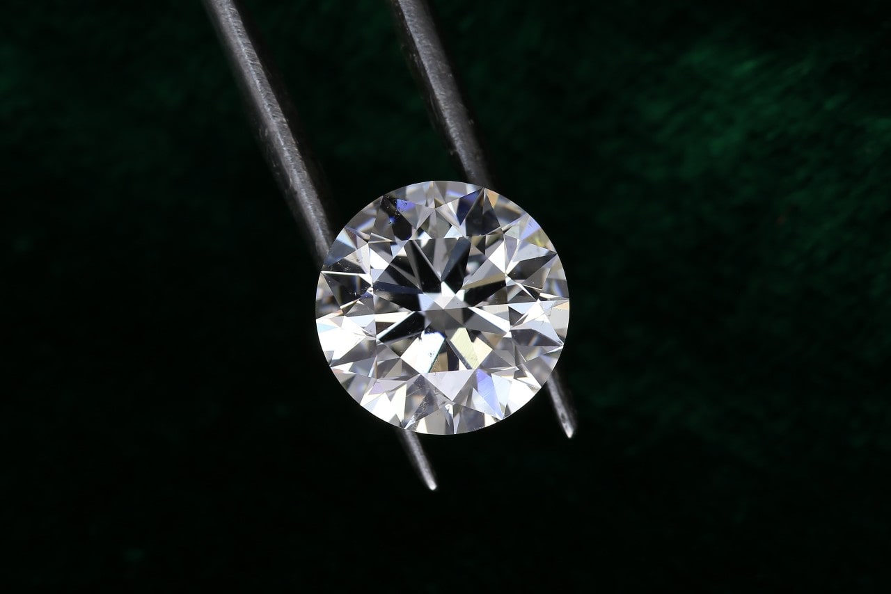 A close-up view of a round brilliant-cut diamond held between metal tweezers against a dark green background.