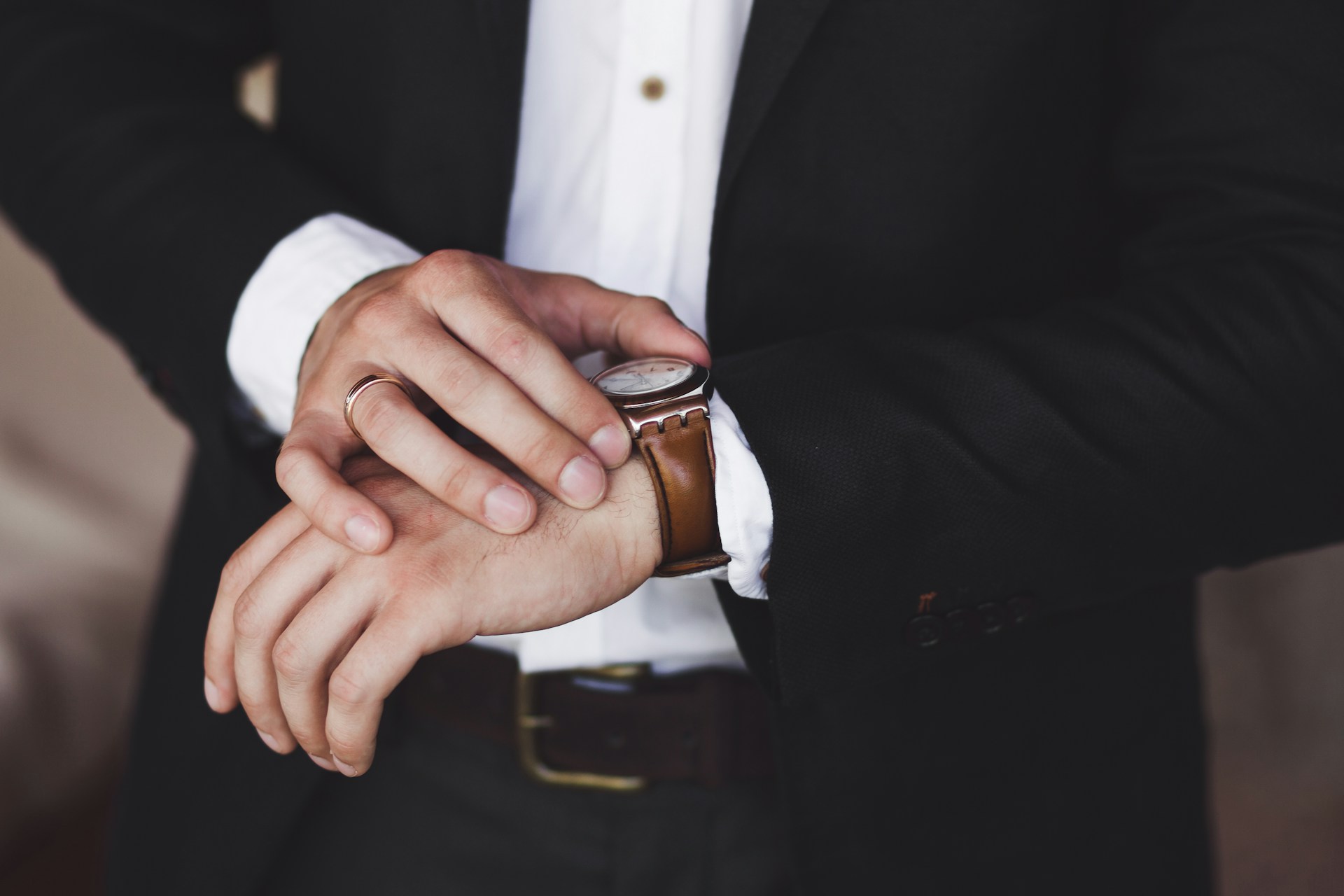 A man in a black suit fixing his brown leather watch with white dial.