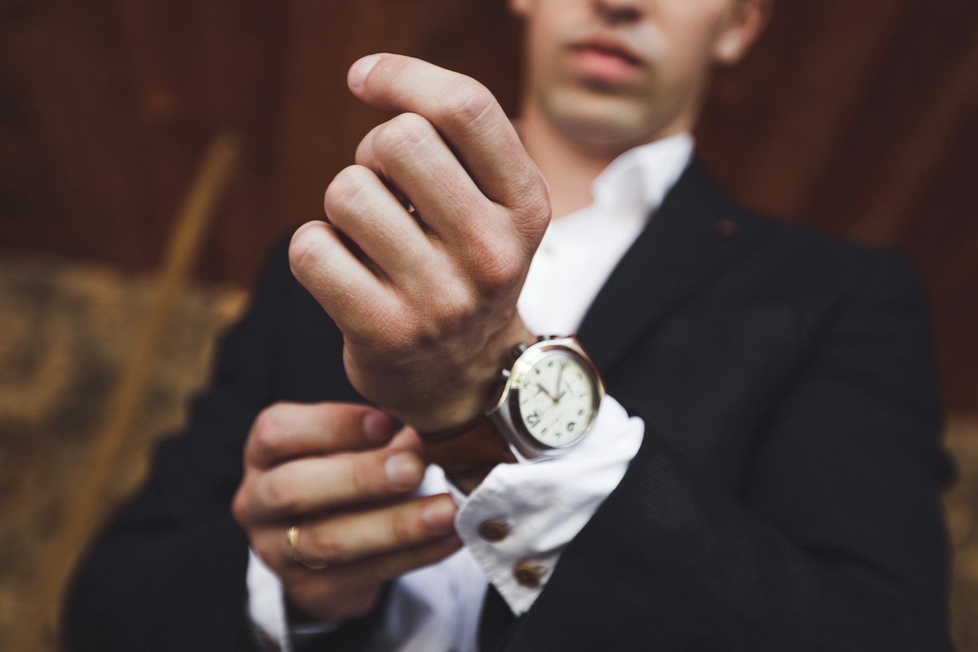 A man in a black suit adjusting his brown leather watch with white dial.