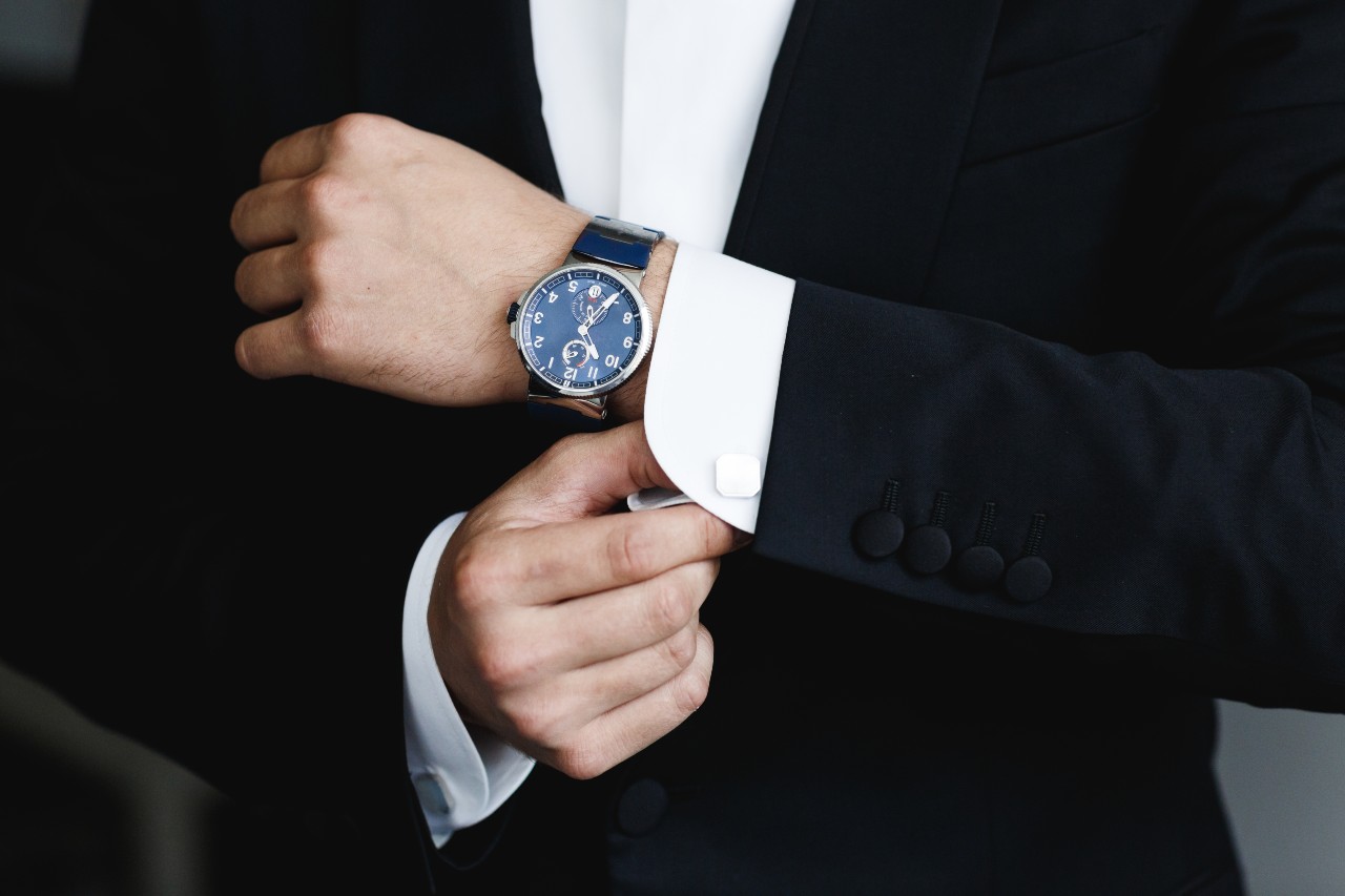 A close up of a man in a black suit fixing his cufflinks, showcasing a black wristwatch with arab numerals and black dial.