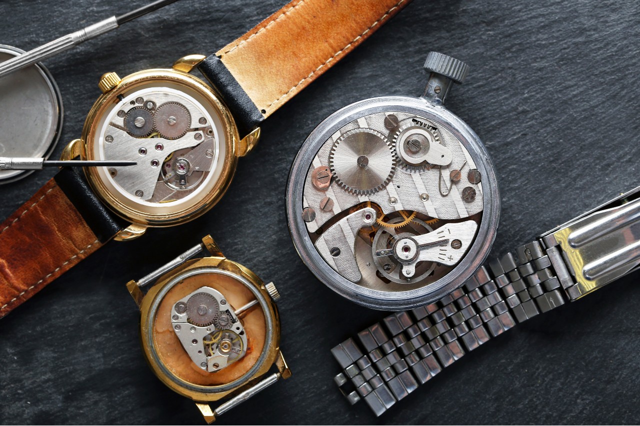 A disassembled mechanical wristwatch with visible gears lies on a wooden surface surrounded by its parts.